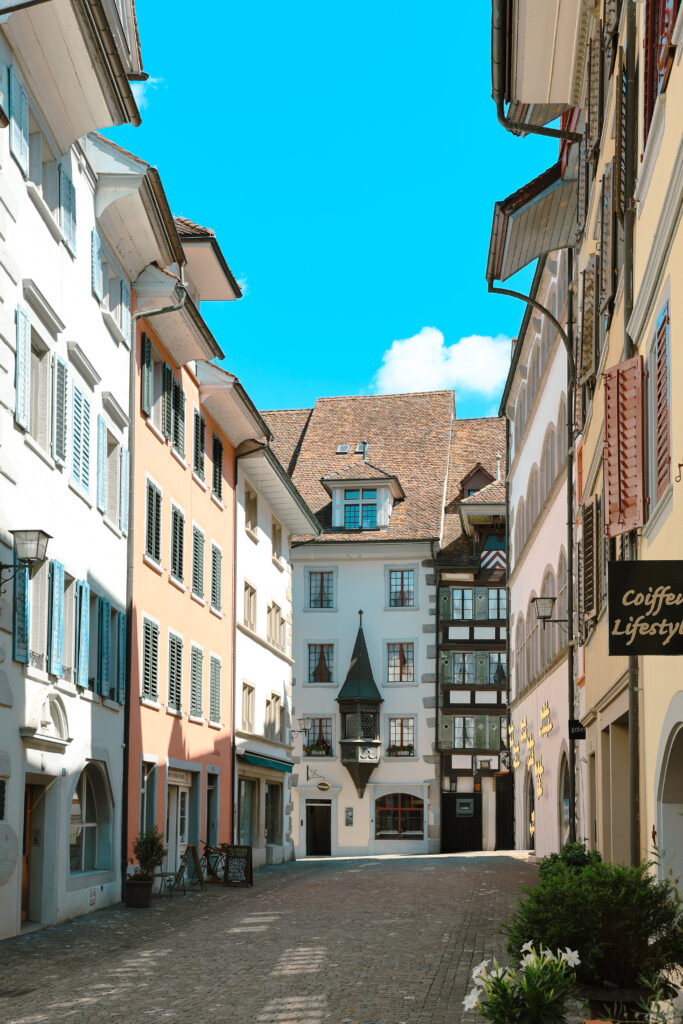 Ruelle ensoleillée de la vieille ville de Zurich, façades pastel beige et pêche, oriel gothique pointu central, ciel bleu vif