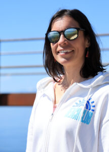 Portrait d'Amandine Devergies souriante, sweat blanc zippé et lunettes de soleil réfléchissantes, au bord du lac Léman à Saint-Saphorin.
