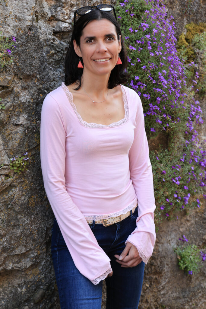 Portrait d'Amandine Devergies souriante, haut rose clair bordé de dentelle, devant un mur de pierre ancienne orné de fleurs violettes à Saint-Saphorin.