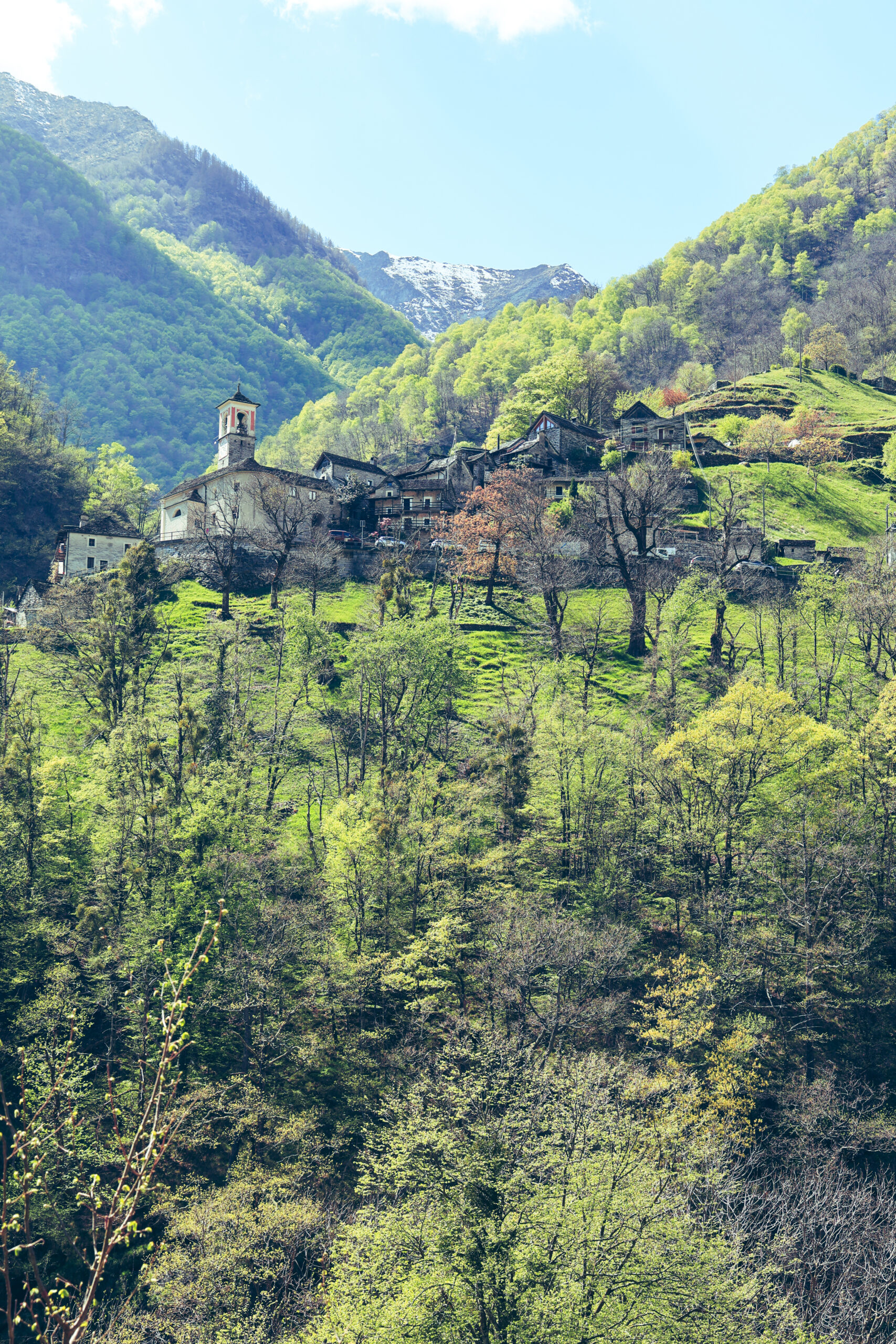 Village tessinois accroché à la montagne avec église et clocher, entouré de forêts verdoyantes, neige en arrière-plan