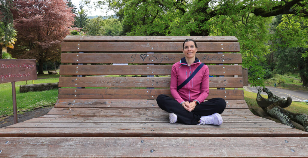 Amandine Devergies assise en tailleur sur un grand banc en bois dans un jardin verdoyant
