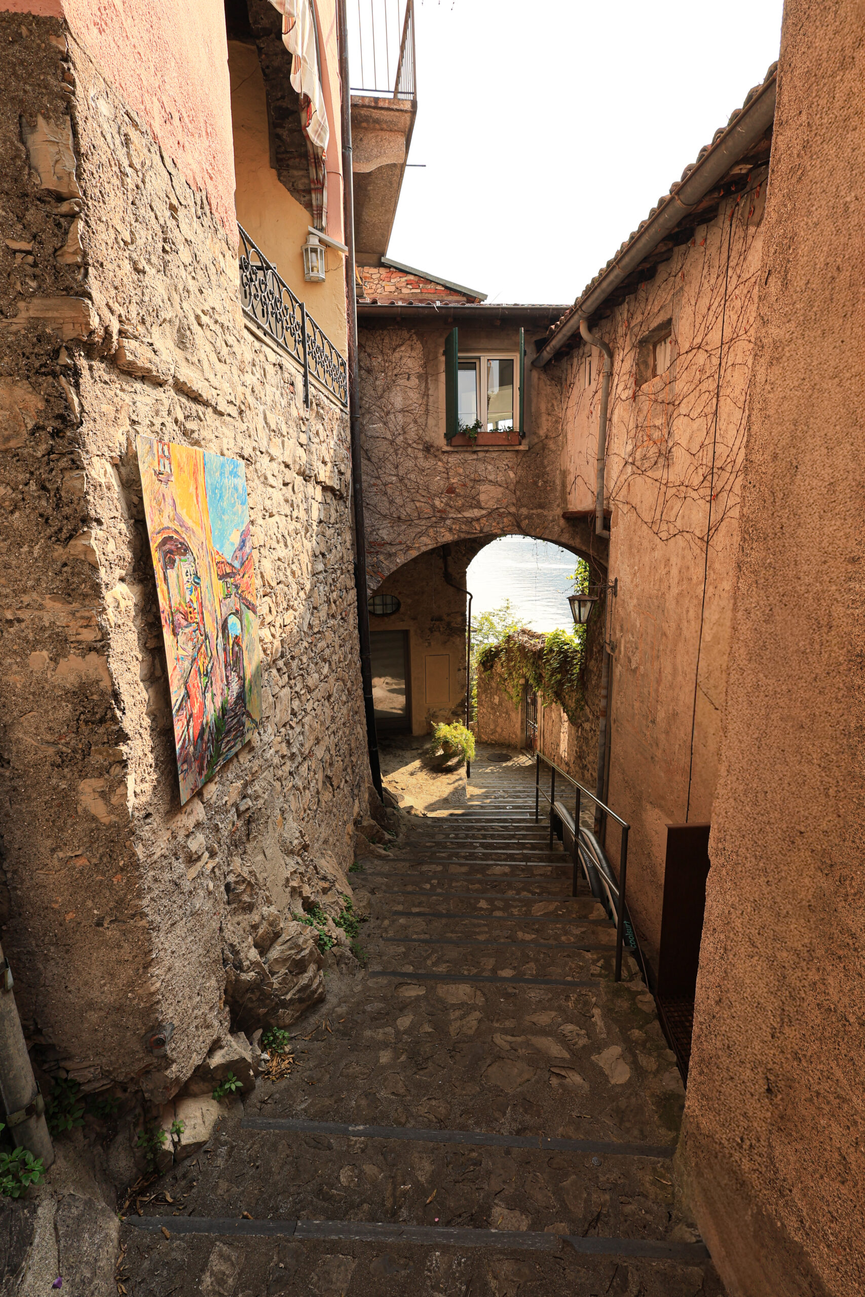 Ruelle en escalier d'un village tessinois avec arc de pierre, fresque colorée et vue sur le lac en fond