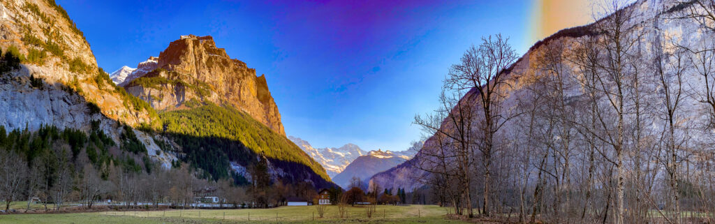 Photographie panoramique de la vallée de Stechelberg dans l'Oberland bernois, montrant des falaises escarpées, des forêts de sapins, des sommets enneigés au loin et un arc-en-ciel naissant sous un ciel bleu.