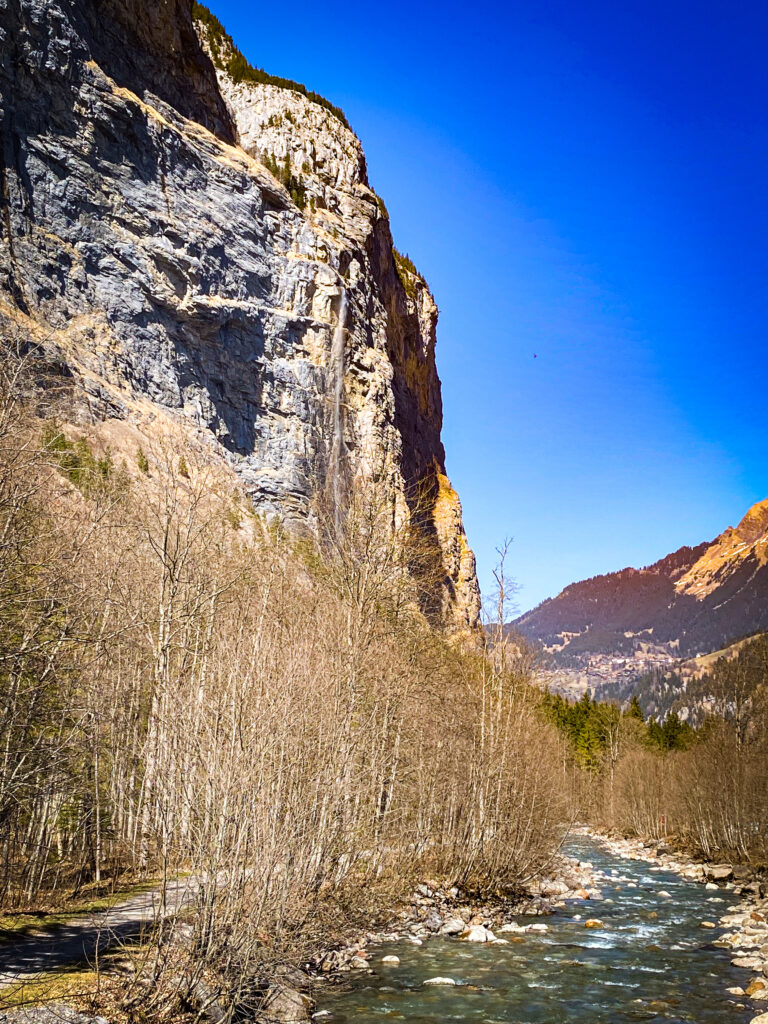 Falaise verticale de la vallée de Lauterbrunnen en hiver, mince cascade sur la roche, rivière et arbres sans feuilles en contrebas, ciel bleu