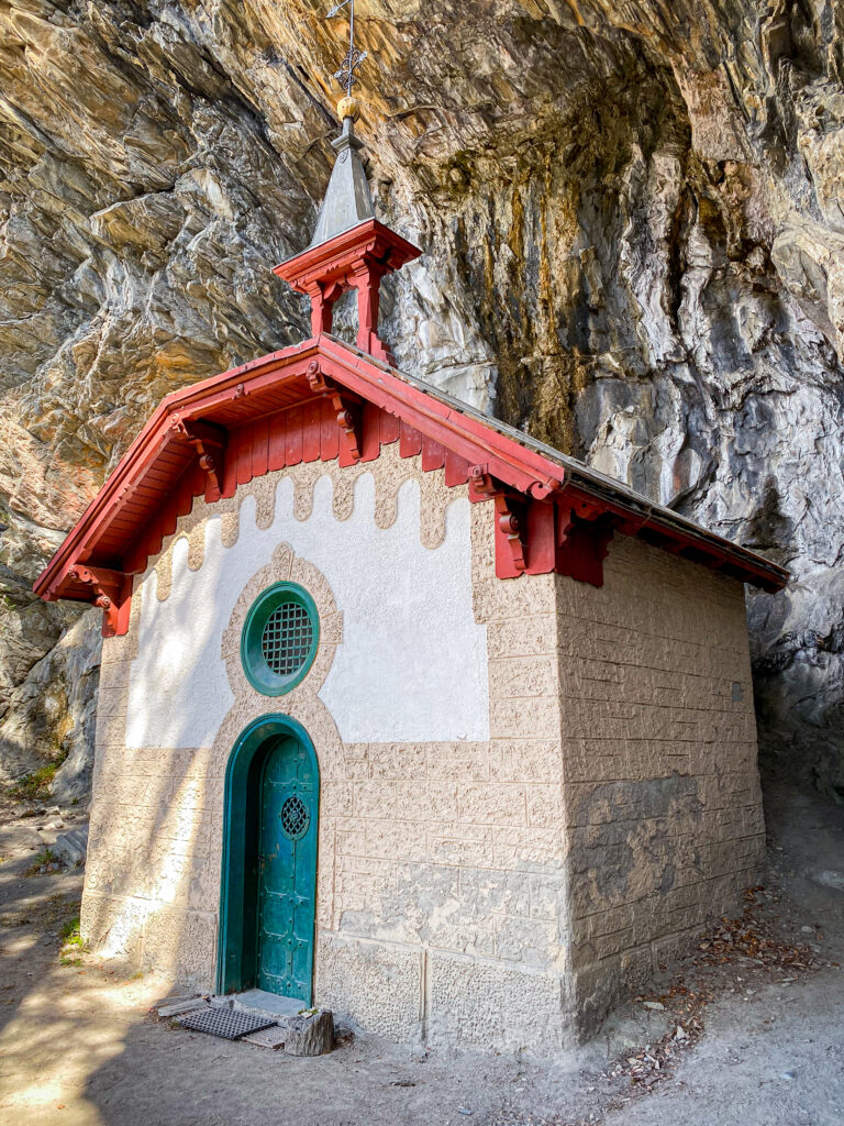 Petite chapelle dans la falaise aux Grisons, toit rouge, porte teal, façade blanche contre la roche
