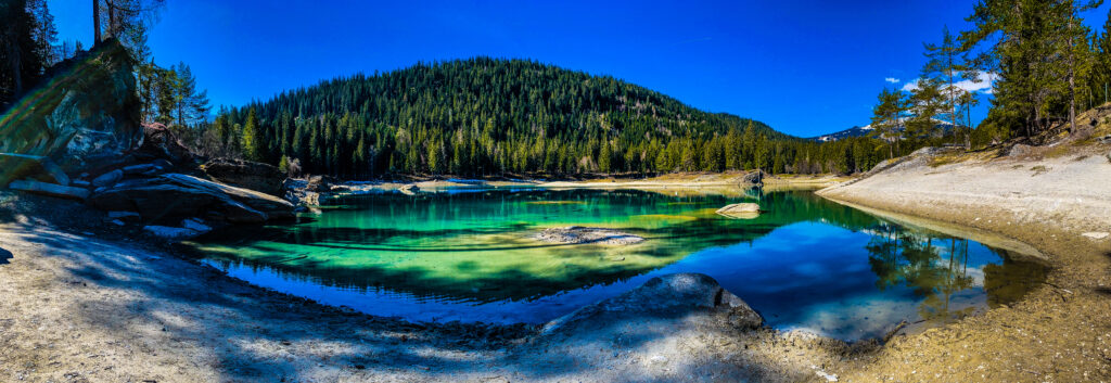 Photographie panoramique d'un lac alpin aux eaux turquoise cristallines à Flims, dans les Grisons (Suisse), reflétant une forêt de conifères sous un ciel bleu intense.