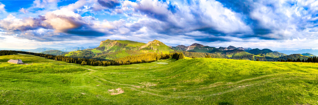 Vaste panorama des alpages suisses verdoyants sous un ciel contrasté de nuages d'orage et de trouées de lumière, avec une petite ferme d'alpage isolée sur la gauche.