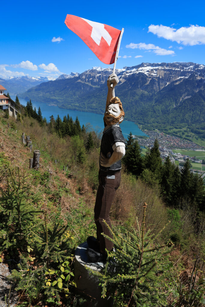 Sculpture en bois d'un personnage brandissant un drapeau suisse face à un lac de montagne