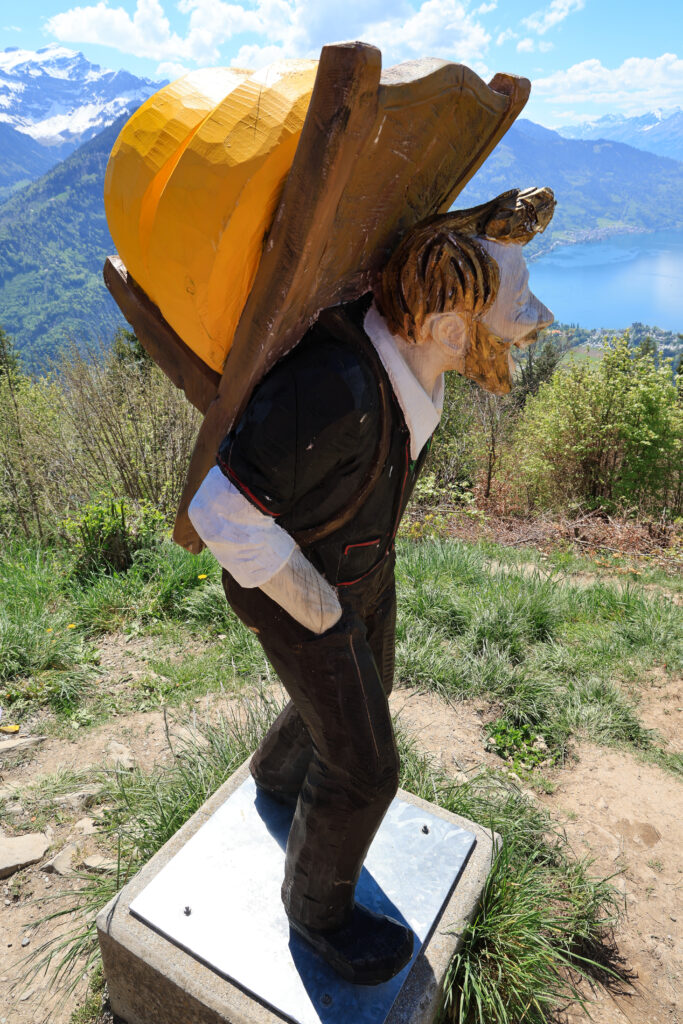 Statue en bois d'un porteur courbé sous une lourde charge, montagnes en arrière-plan